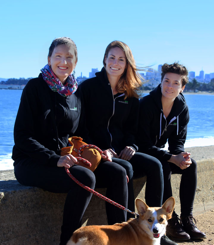 Three smiling women sitting in front of the San Francisco Bay with a Corgi dog on leash.