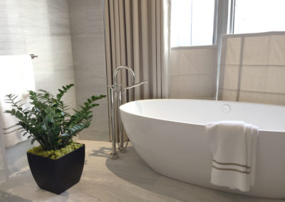 A modern bathroom with a ZZ plant in a matte black container, next to a white oval bathtub in front of big windows.